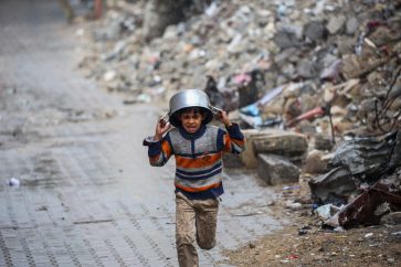 TOPSHOT - A displaced boy covers his head with a pan as he runs from the rain past building rubble at the Bureij refugee camp in the central Gaza Strip on November 24, 2024, amid the ongoing war between Israel and the Palestinian Hamas militant group. (Photo by Eyad BABA / AFP)